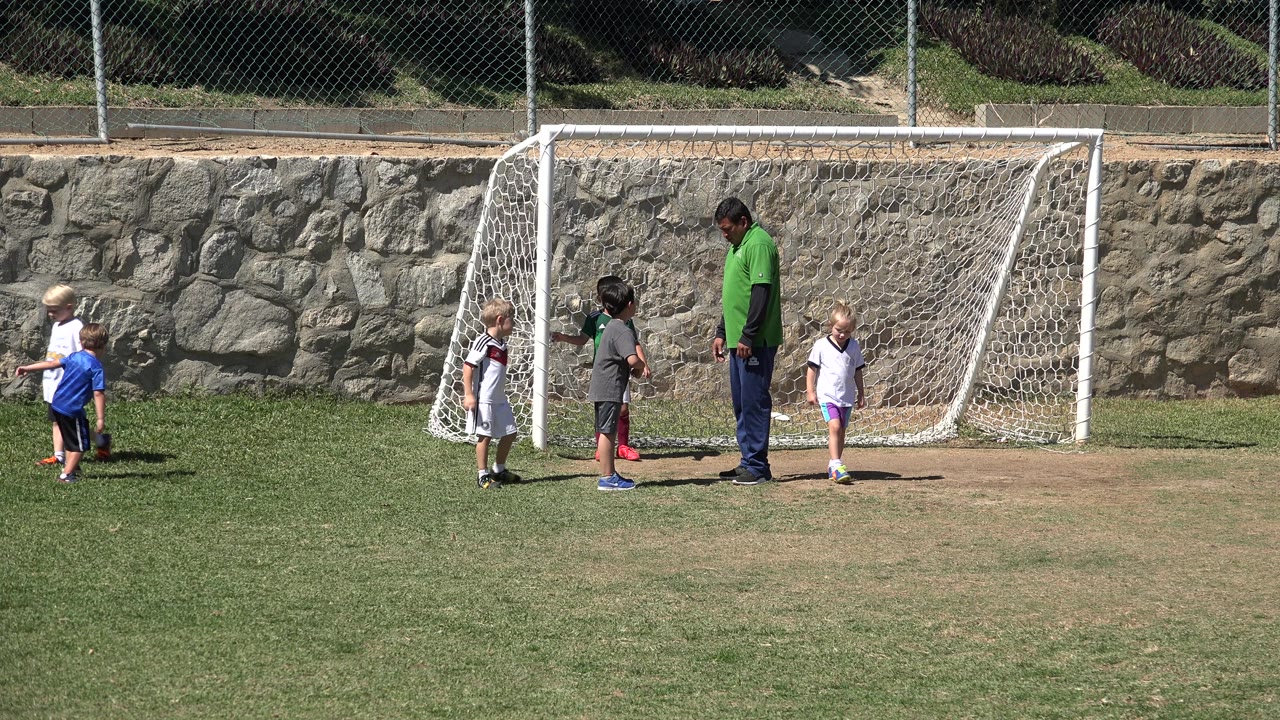 Children remain spread around the goal area in San Jose del Cabo, Mexico, with one child in blue at left and the adult guide standing near the center.