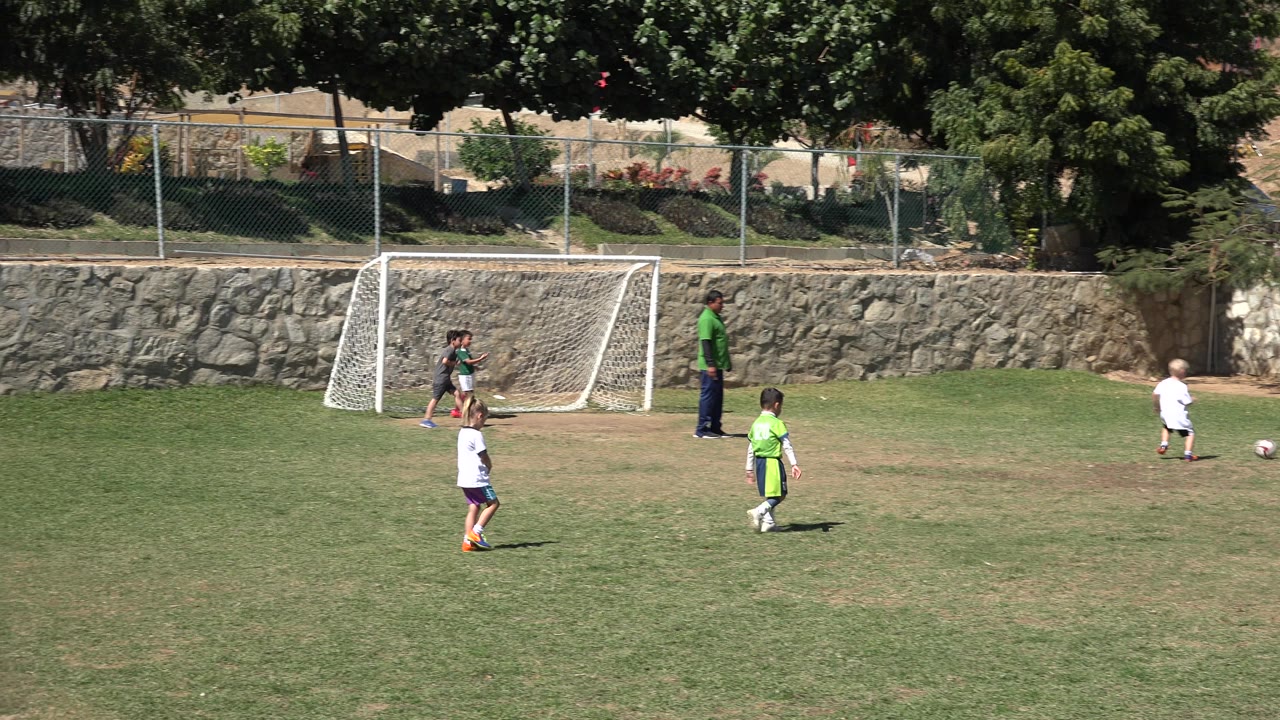 Children stand and move near a white soccer goal on a grass field in San Jose del Cabo, Mexico, with an adult supervising from the midground.