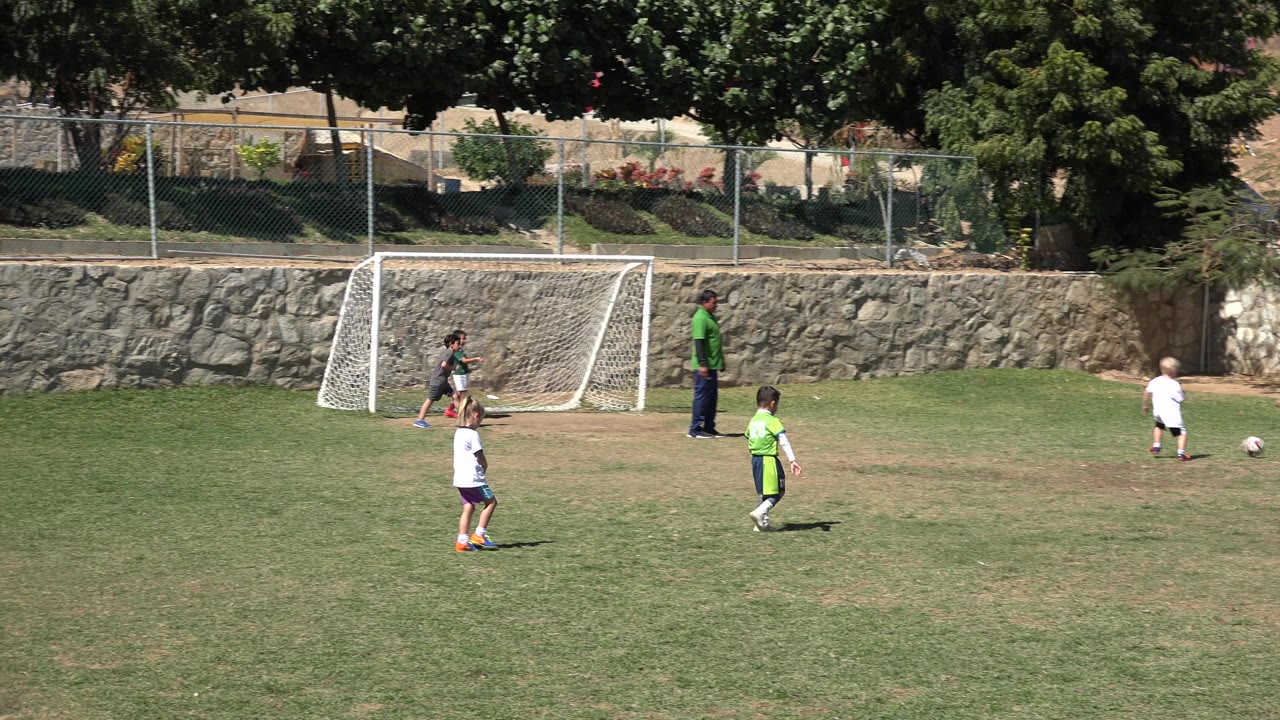 Children spread out across the grass near the white goal in San Jose del Cabo, Mexico, while the ball sits in the open field and an adult watches nearby.