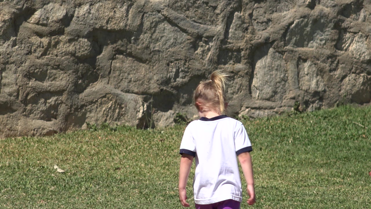 At 0:01, the child remains turned away in the grassy midground of San Jose del Cabo, Mexico, with the rough stone wall occupying the upper frame.