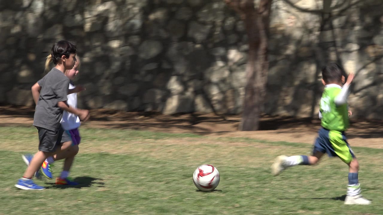 Two children in athletic clothes chase a red and white soccer ball across grass in San Jose Del Cabo, Mexico, with the shaded wall and trees behind them.