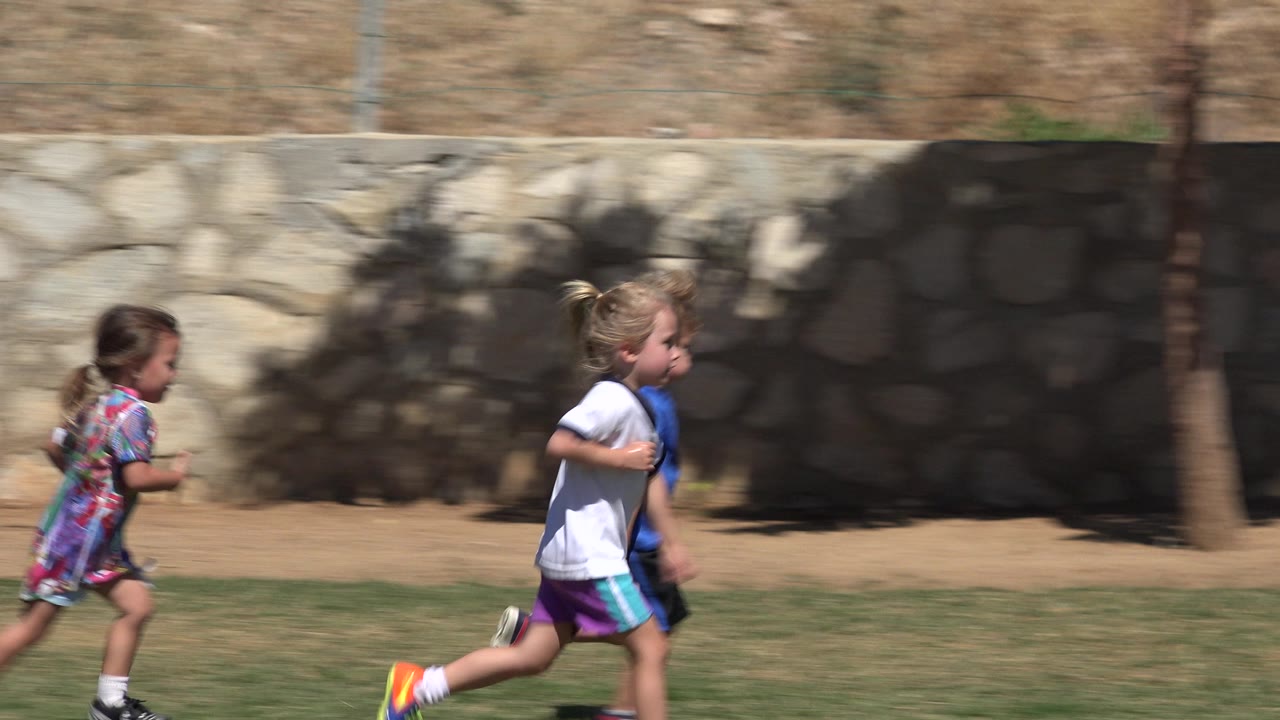 A girl in a patterned dress and two children in athletic shirts run across the grass in San Jose Del Cabo, Mexico, with the wall filling the background.