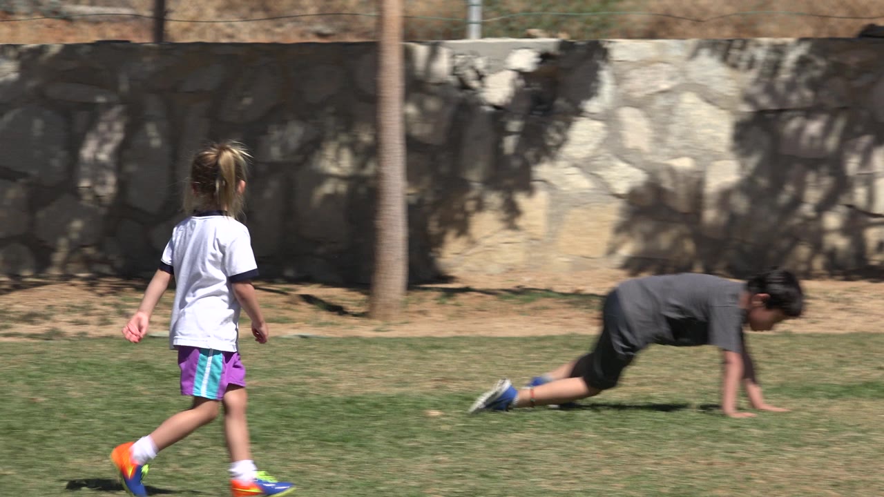Girl runs across a grass soccer field while another child is low to the ground.