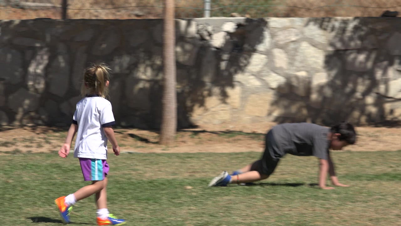 A child in a white shirt and purple shorts runs through the sunlit field in San Jose Del Cabo, Mexico, while another child is down on hands and feet near the wall.