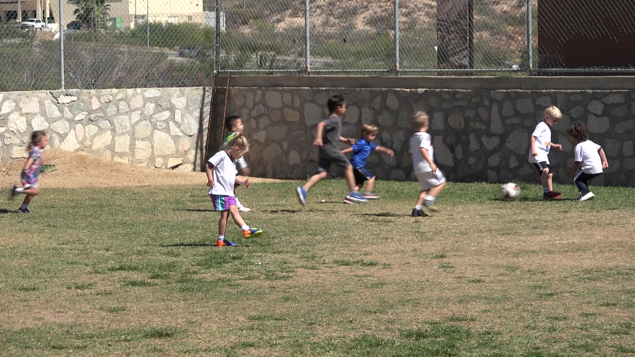 Several children move toward a soccer ball on the grass in San Jose del Cabo, Mexico, while a girl in a white shirt stands nearer the left foreground.