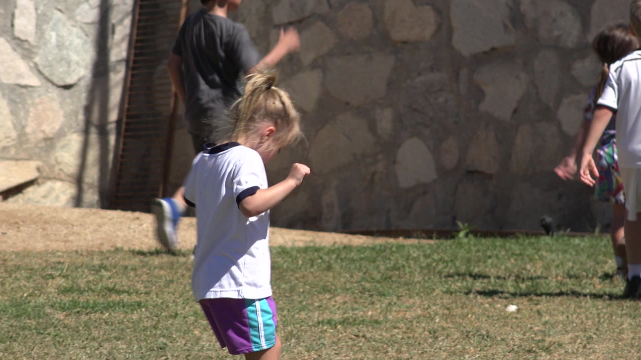 At 0:01 in San Jose del Cabo, Mexico, the young girl continues across the grass with her head down and children partly visible behind her.