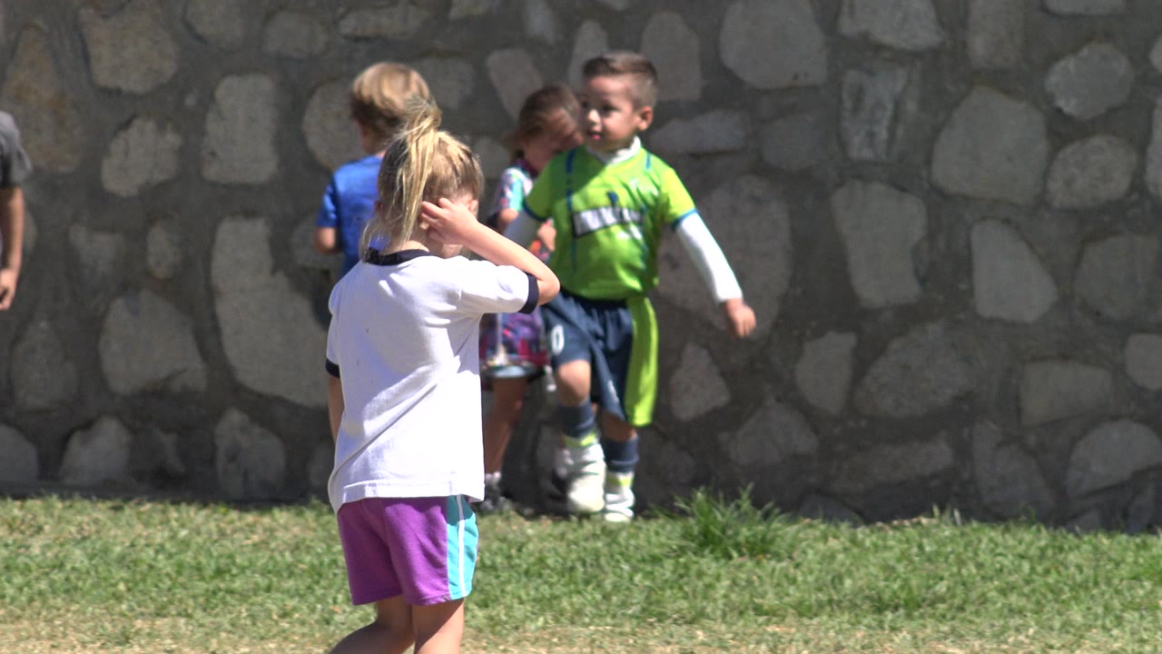 The girl in the white shirt moves across the foreground as children in green and blue soccer outfits remain near the stone wall in San Jose del Cabo, Mexico.