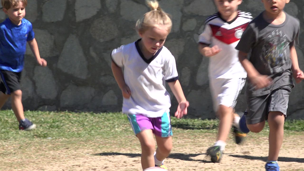 Children run toward the camera on a patchy grass soccer field in San Jose Del Cabo, Mexico, with a stone wall behind them.