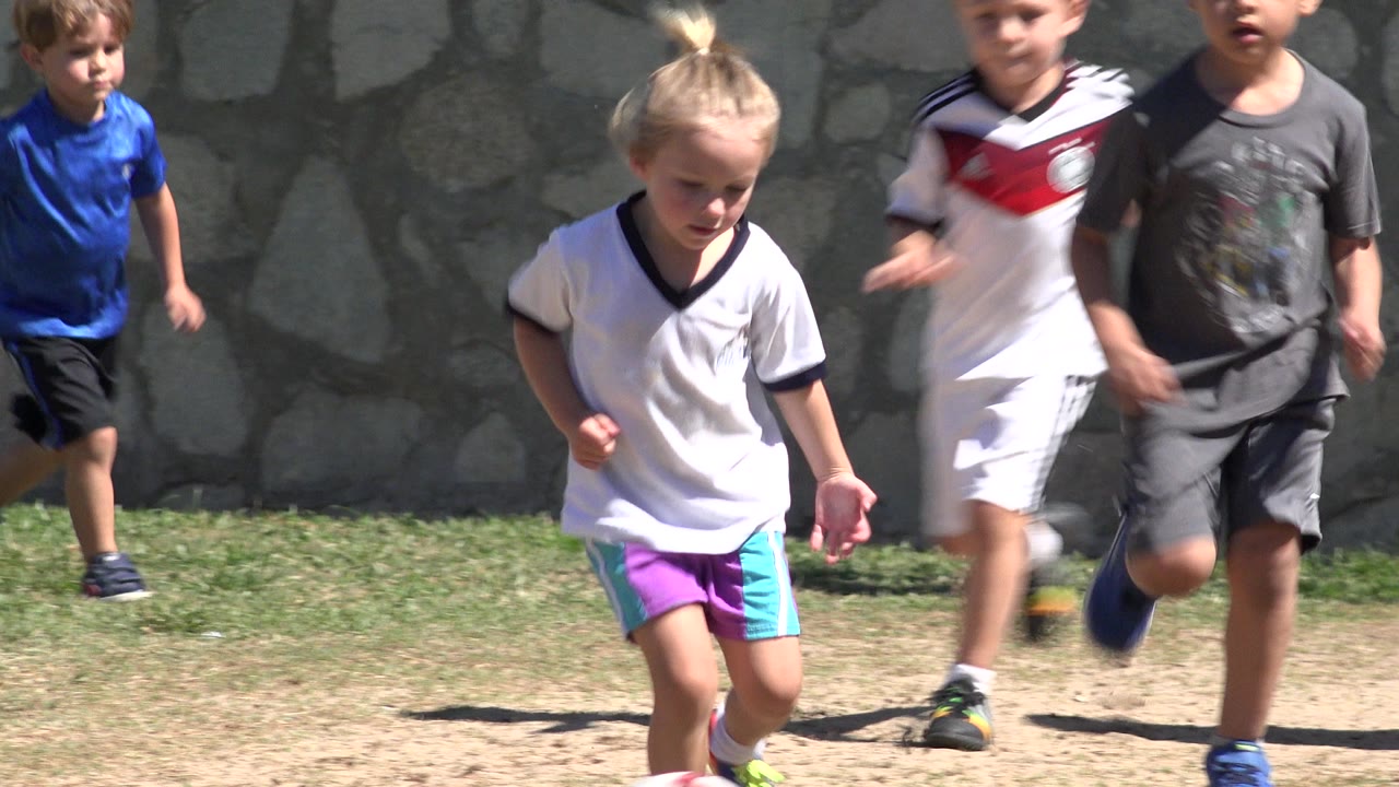 Several children run in a tight group across the grass in San Jose Del Cabo, Mexico, with bright sun and the stone boundary wall behind them.