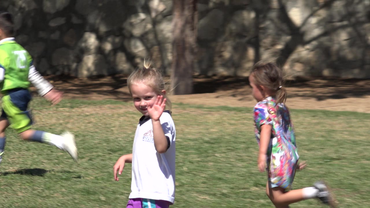 A blonde young girl in a white shirt waves toward the camera on a grass field in San Jose del Cabo, Mexico, as other children run past behind her.