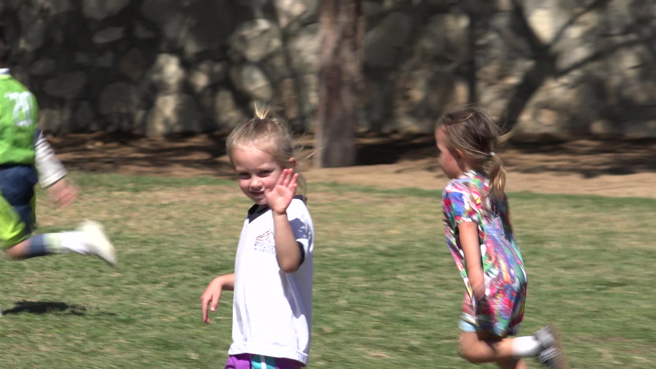 The girl faces the camera with her hand raised while a child in green sports clothing runs behind her across the grass in San Jose del Cabo, Mexico.