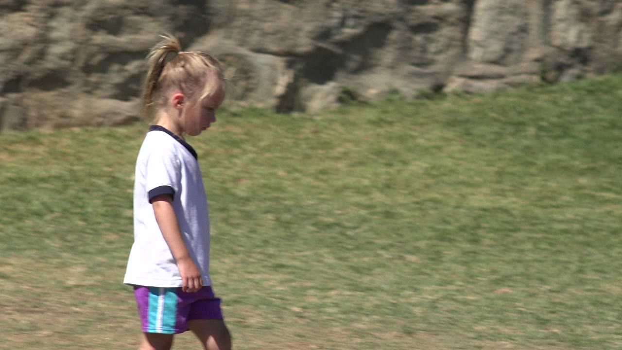 A young blonde girl in a white shirt and purple shorts walks left to right across a grass field in San Jose del Cabo, Mexico, with a stone wall behind her.