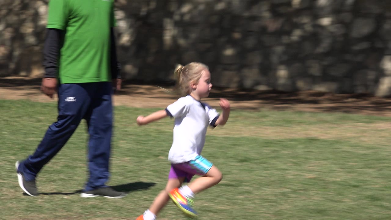 The child continues running across the grass in San Jose del Cabo, Mexico, with the adult's legs and green shirt still visible near the left side.