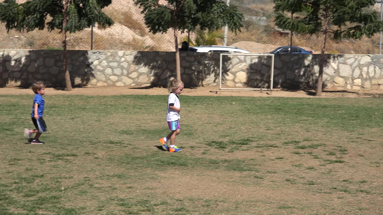 Two children stand and move on a sunlit soccer field in San Jose del Cabo, Mexico, with a colorful ball near the child in white and a stone wall behind them.
