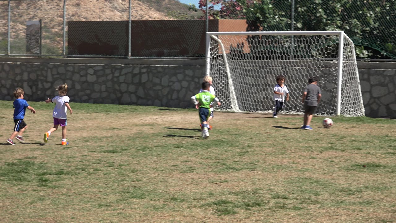 Children run toward a soccer goal on a dry grass field.