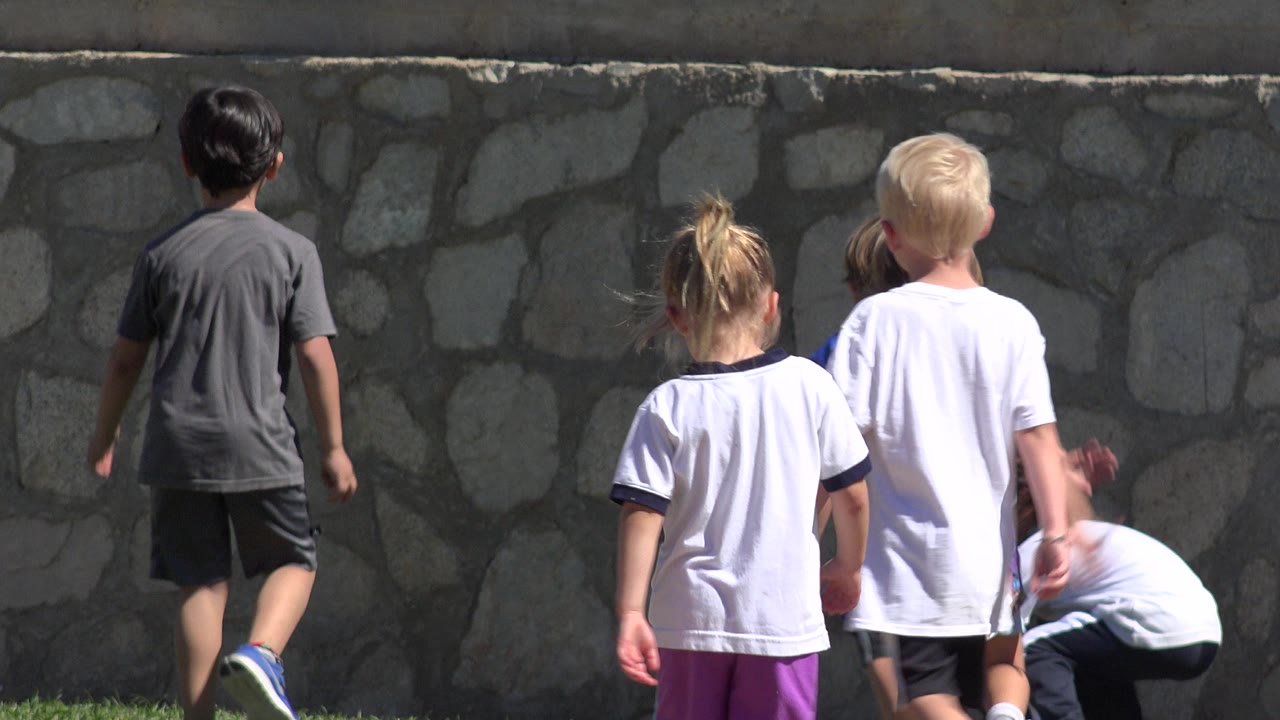 Children in athletic clothing walk away from the camera on a grass field in San Jose del Cabo, Mexico, with a gray stone wall behind them.