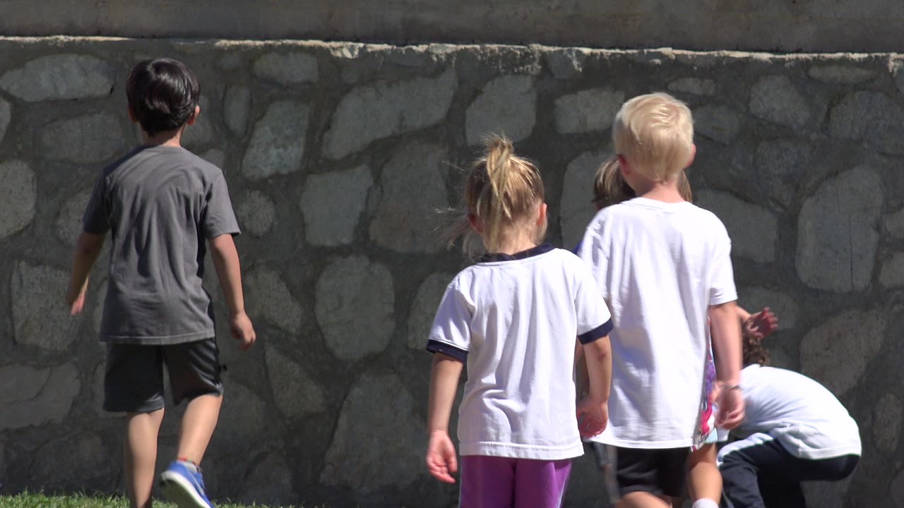 Several children stand and bend near a stone wall on the grass field in San Jose del Cabo, Mexico, with bright sun on white shirts and purple shorts.