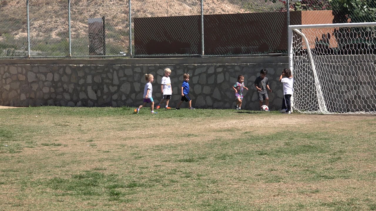 Wide frame of children moving along the goal area in San Jose Del Cabo, Mexico, with the red and white soccer ball near the right side of the group.