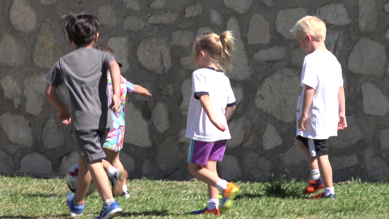 Children walk and run near a soccer ball beside a stone wall.