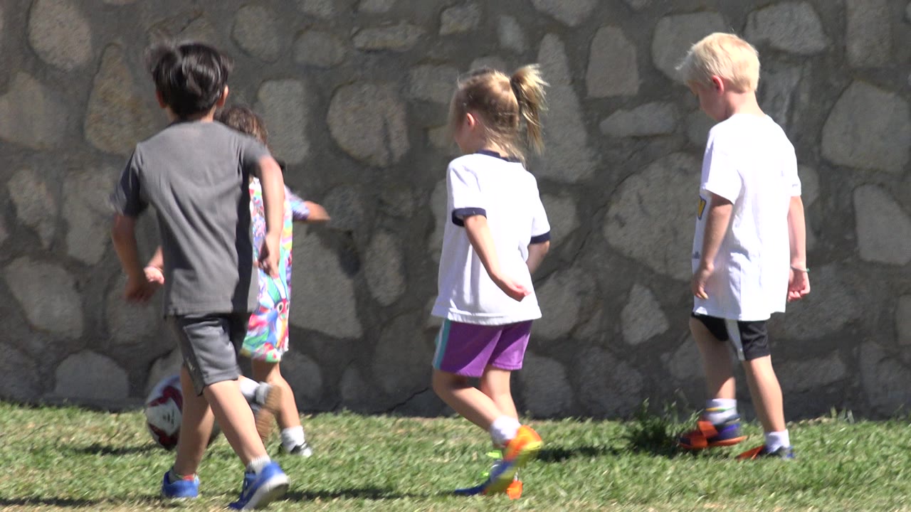 Three children continue along the grass in San Jose del Cabo, Mexico, with the soccer ball low in frame near the stone wall and strong sunlight on their shirts.