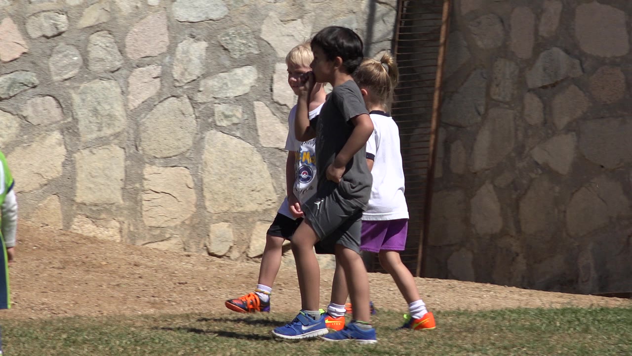 Children in sportswear walk past a stone wall in San Jose Del Cabo, Mexico, with dirt and grass at their feet.