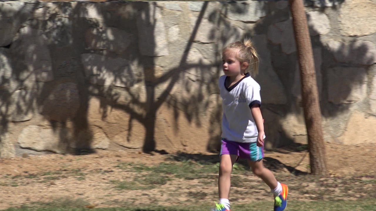 The girl continues along the wall in San Jose del Cabo, Mexico, with one foot forward, bright sneakers visible, and dark tree shadows spread across the stone behind her.