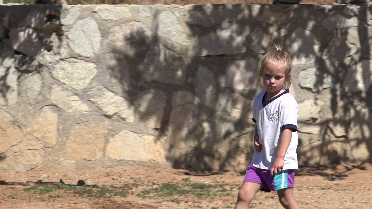 The child continues walking in San Jose del Cabo, Mexico, wearing a loose white shirt and purple shorts against a pale stone wall with hard shadows.