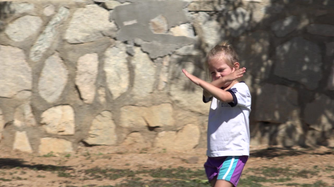 The girl remains near the center of frame in San Jose Del Cabo, Mexico, with her arms crossed upward and sunlit stonework behind her.