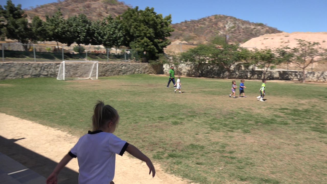 The girl begins moving out from the shaded sideline toward children gathered on the sunlit soccer field in San Jose del Cabo, Mexico.