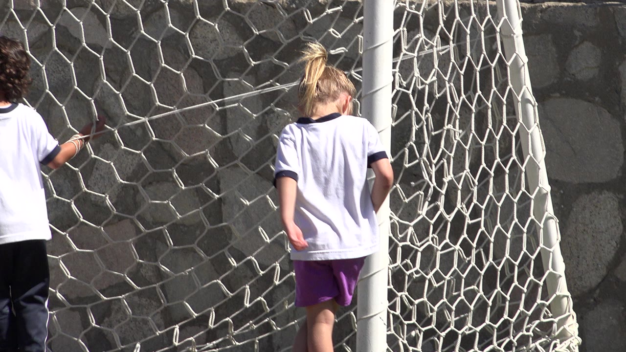 Young girl walking beside a soccer net near a stone wall.