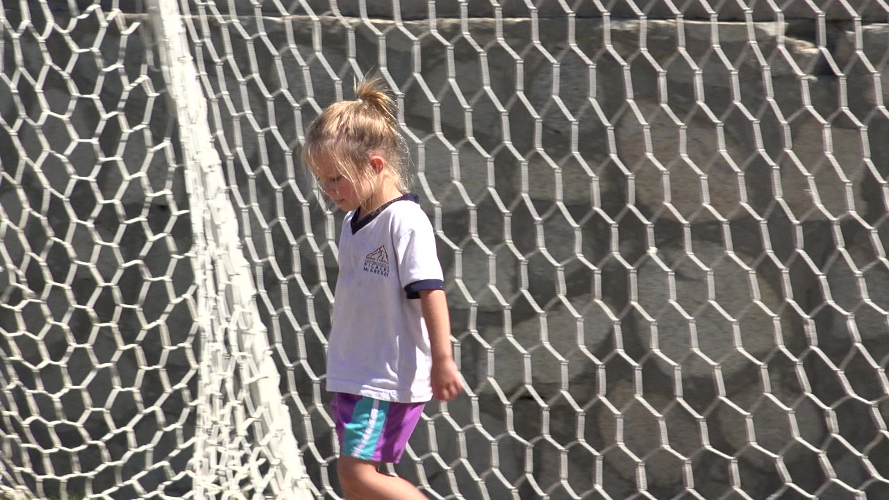 The girl continues across the frame in front of the sagging white soccer net, with the stone wall filling the background.