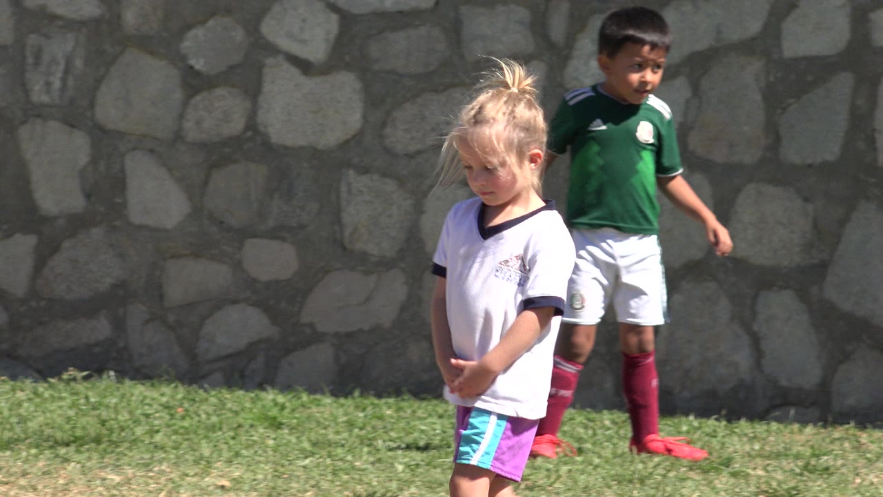 The girl remains in the foreground on the grass while a boy in green and white soccer clothing moves behind her against the stone wall in San Jose del Cabo, Mexico.