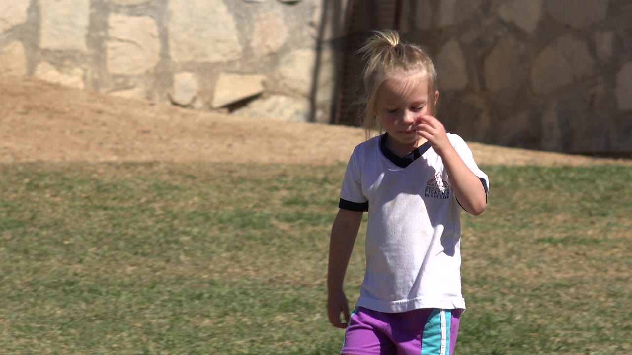 The girl continues across the San Jose del Cabo soccer field while touching her face, with patchy grass and a pale stone wall in the background.
