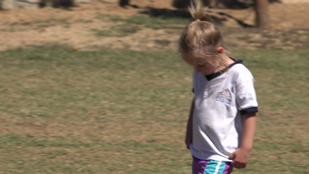Blond child walking on grass with her head lowered.