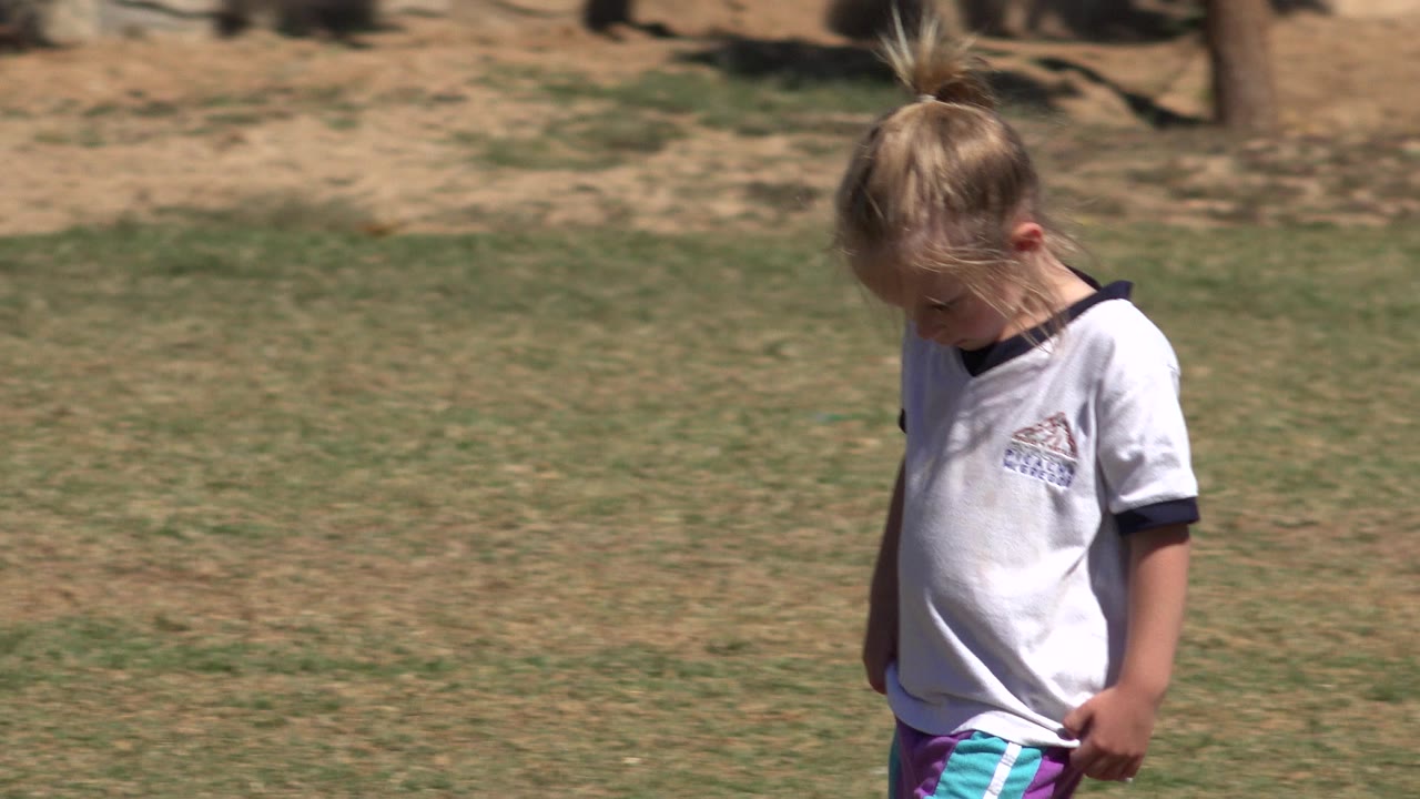 The girl continues through the grassy park in San Jose del Cabo, Mexico, with her head tilted down and her white shirt bright in direct sun.