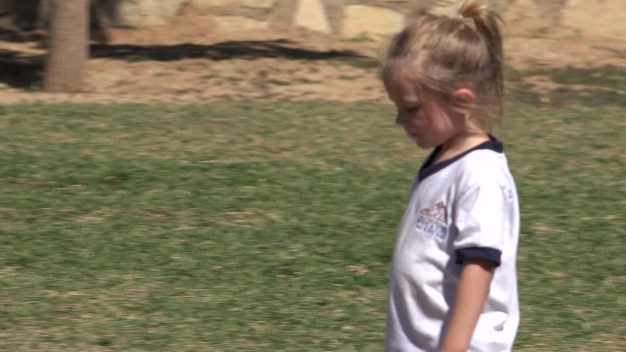 Young girl in a white jersey walking across grass in profile.