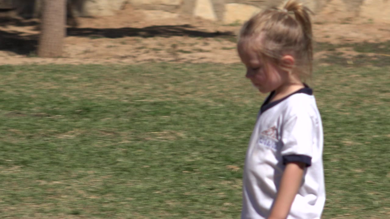 The girl continues walking across the San Jose del Cabo grass field, with her white jersey bright in direct sun and the background slightly soft.