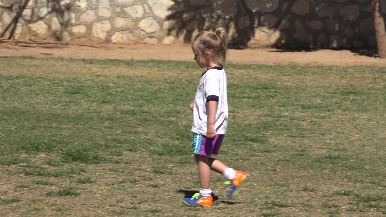 At 0:00 in San Jose Del Cabo, Mexico, a young girl in a white shirt and purple shorts walks left across patchy grass with trees and a stone wall behind her.