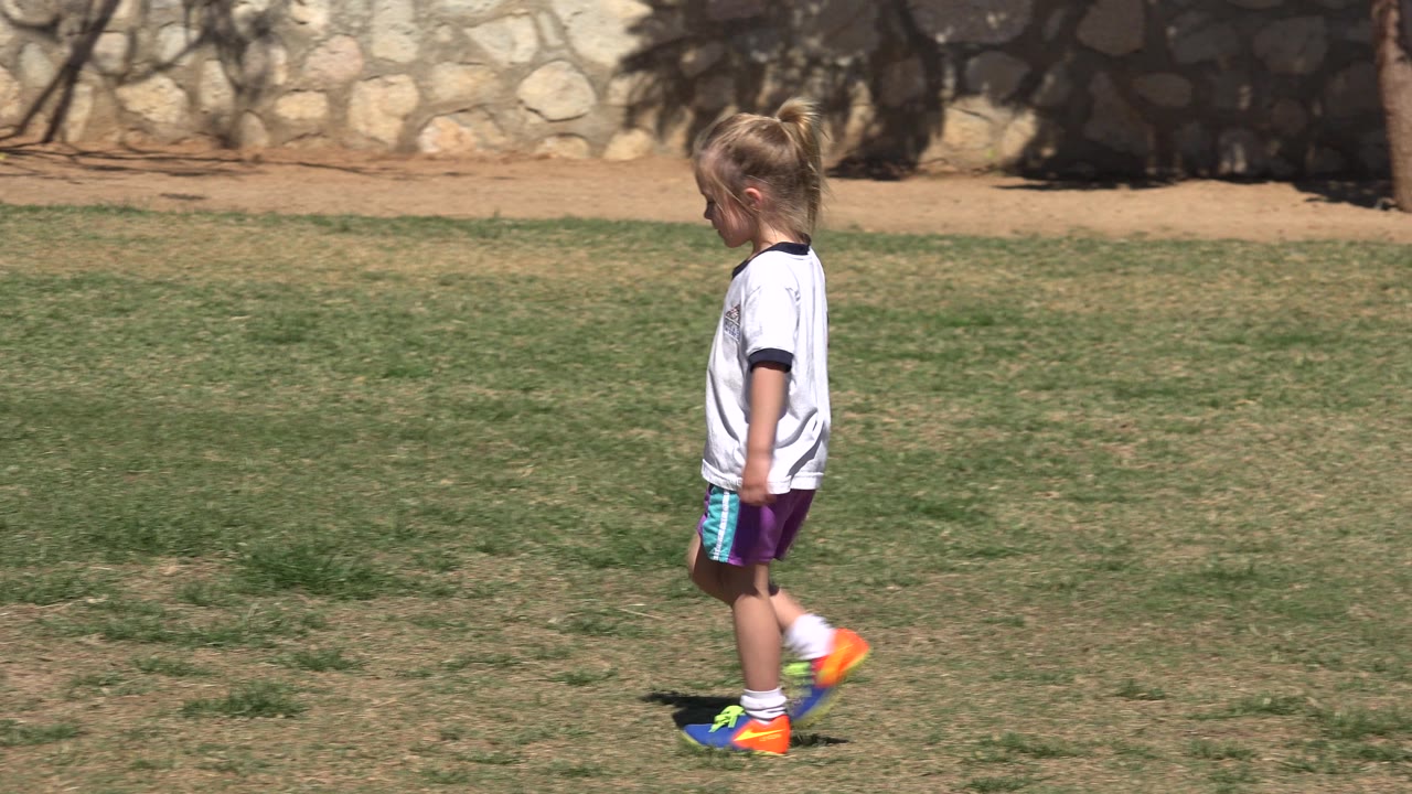 At 0:01 in San Jose Del Cabo, Mexico, the girl continues moving left through the open grass, with her head angled downward and the stone wall still behind her.