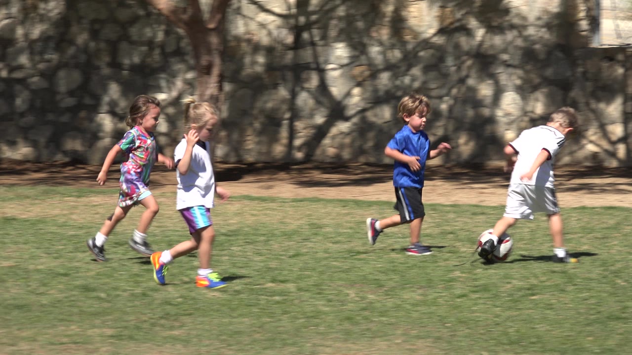 Children continue running across the grass in San Jose del Cabo, Mexico, with the boy in white closest to the soccer ball and tree shadows on the wall.