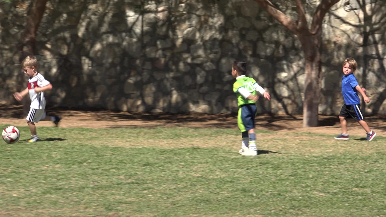 Children play soccer on a grass field in San Jose del Cabo, with a red and white ball in the foreground and a stone wall and trees behind them.