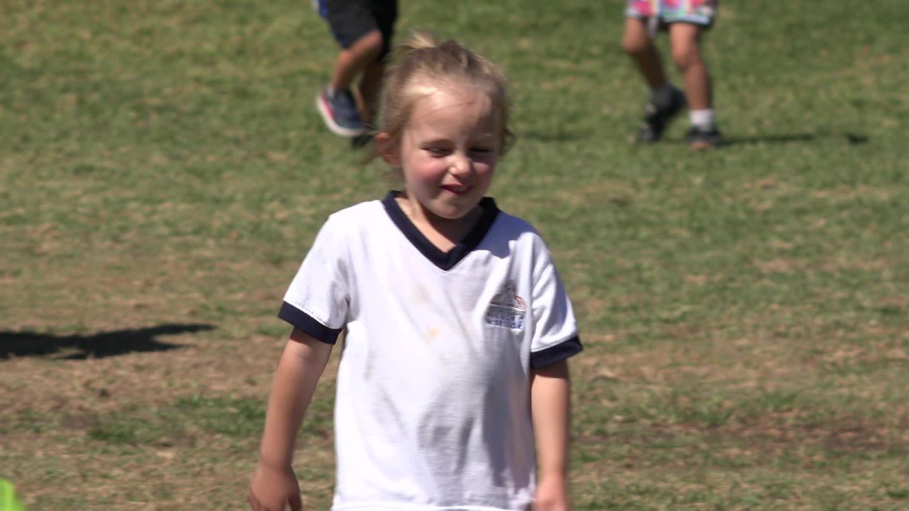 Young girl in a white sports shirt stands on a dry grass field during outdoor soccer practice in San Jose del Cabo, Mexico, with partial figures in the background.