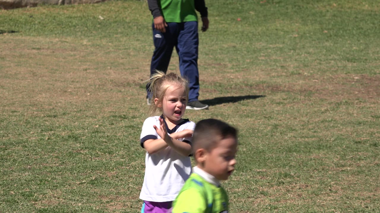 Young girl in a white soccer jersey gesturing on a grass field as a boy passes in front.