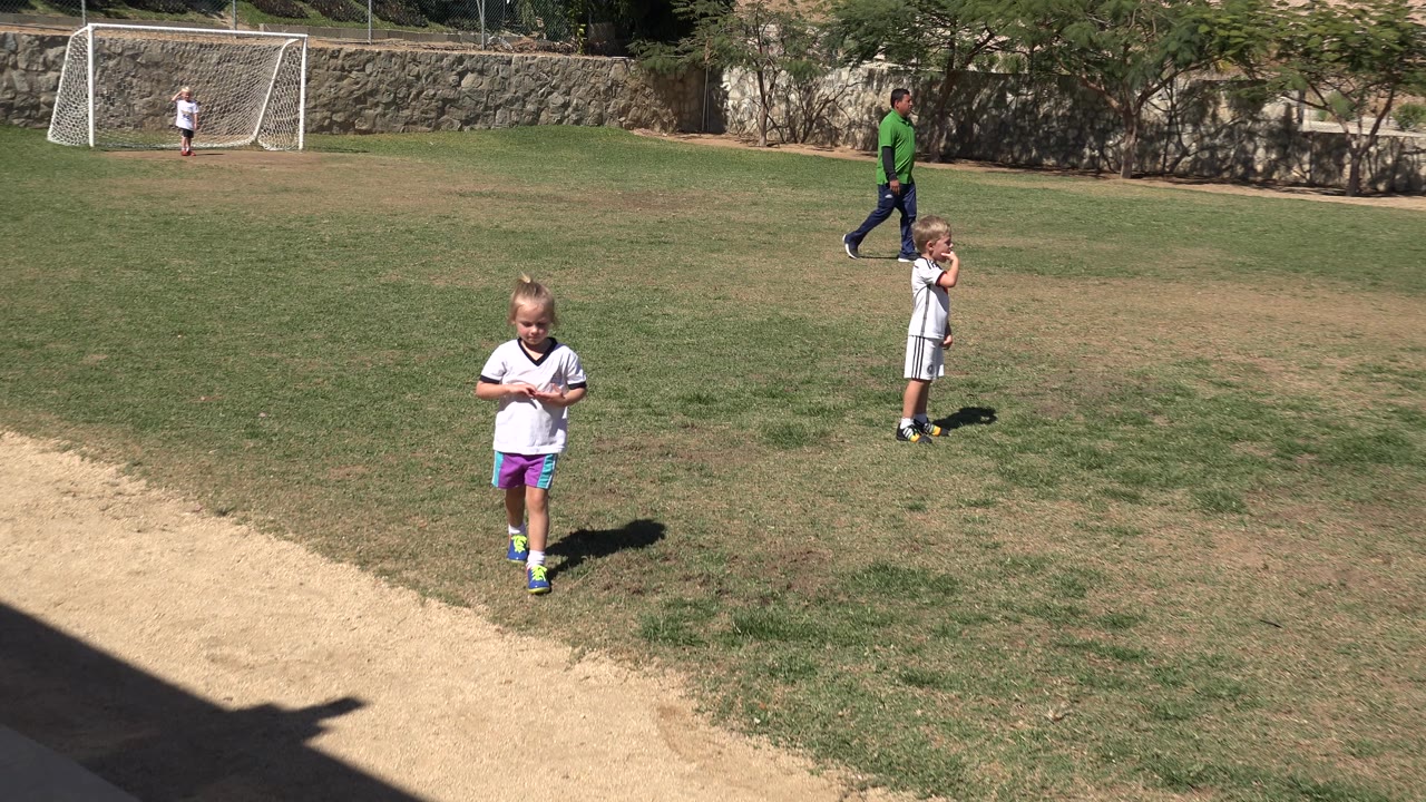 A young girl in a white soccer shirt stands near the camera on a grass and dirt soccer field in San Jose Del Cabo, Mexico, with another child, a coach, and a goal net behind her.