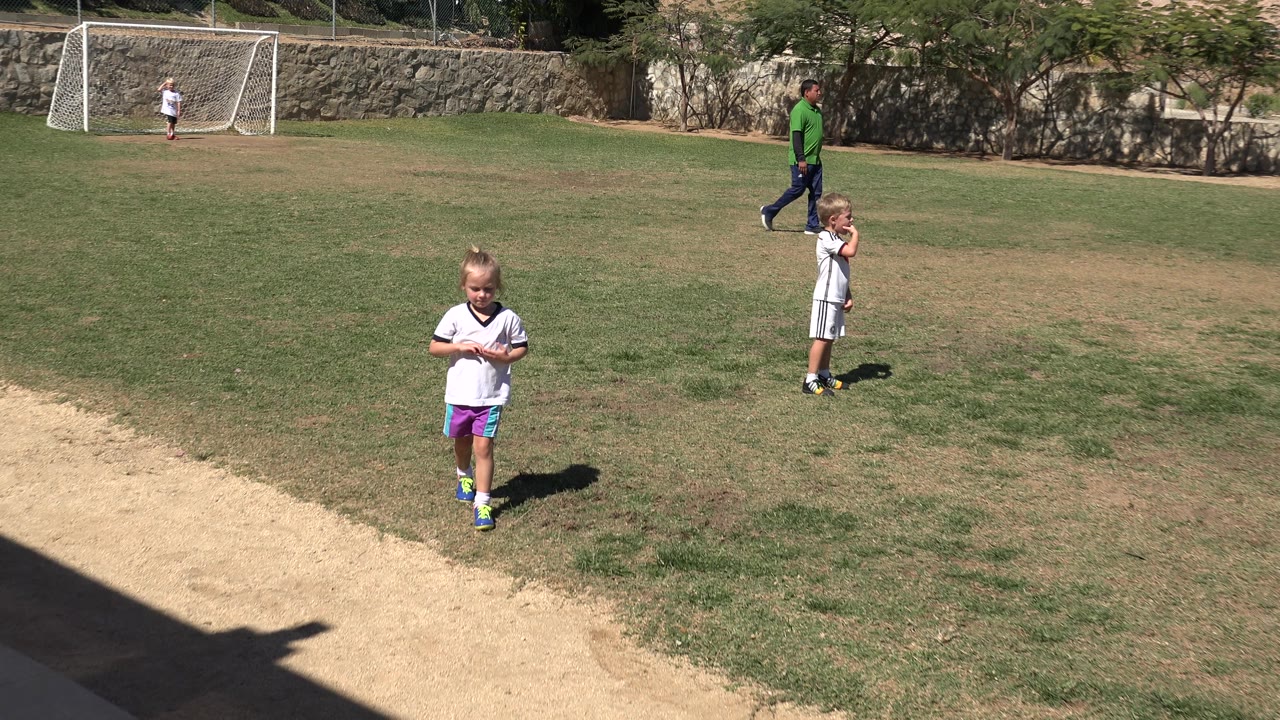 The girl remains near the center foreground on the San Jose Del Cabo soccer field as a child stands farther back and an adult in a green shirt walks near the goal area.