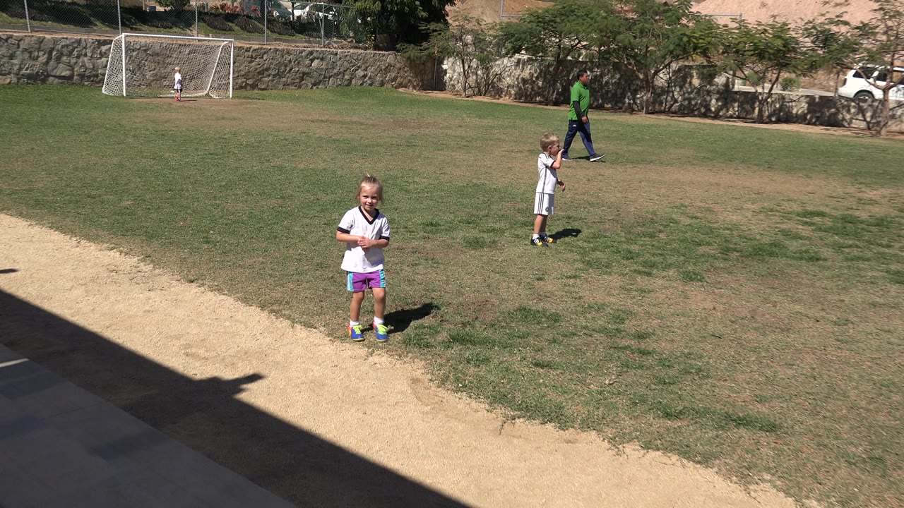 A young girl stands farther from the camera on the sunlit field in San Jose Del Cabo, Mexico, while another child and an adult appear near the trees and a goal net in the background.