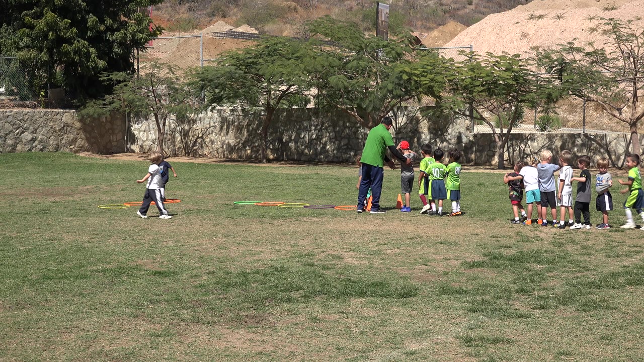 In San Jose del Cabo, Mexico, a coach in a green shirt faces a cluster of children as another child crosses the left side near the training hoops.