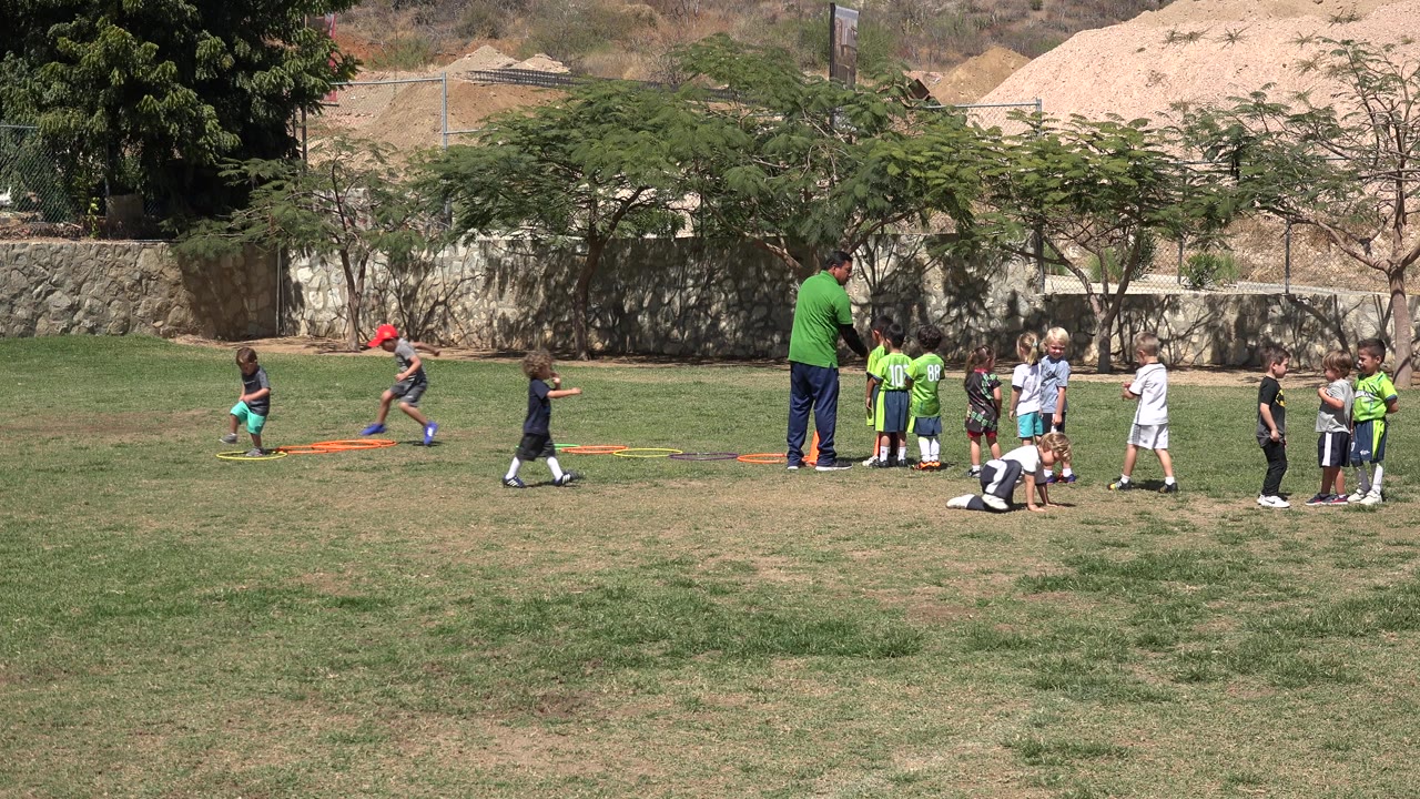 Children gather around a coach on a sunlit soccer field in San Jose del Cabo, Mexico, while a few kids run near colored training hoops.