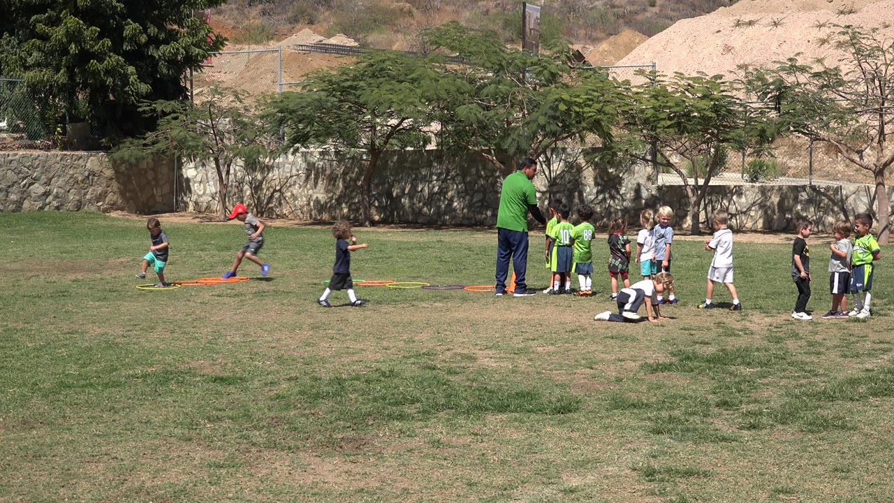 A child in a red cap runs near orange hoops while the coach stands with a group of children on the field in San Jose del Cabo, Mexico.