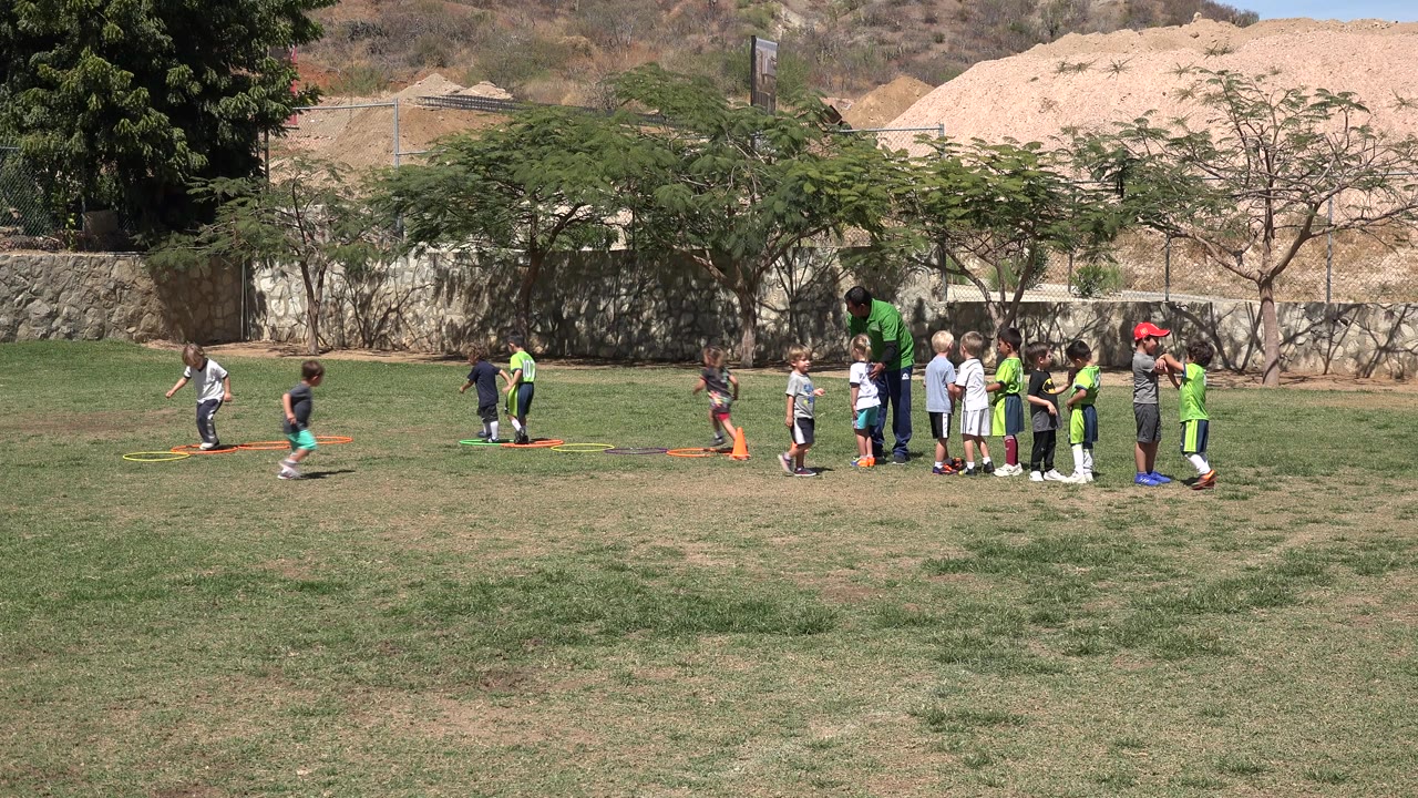 A boy runs across the grass near colored hoops while a line of children and supervising adults stand to the right in San Jose Del Cabo, Mexico.